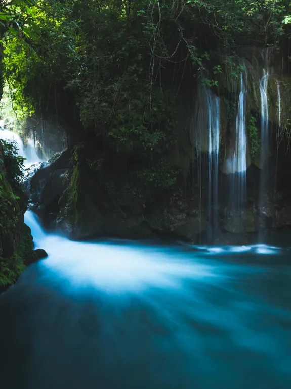 A serene waterfall cascades into a turquoise pool surrounded by lush green foliage. The water appears silky and smooth due to a long exposure effect. Sunlight filters through the dense canopy, creating a tranquil and secluded atmosphere.