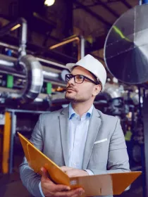 A person wearing a white hard hat and glasses holds a folder while standing in an industrial facility with large metal pipes and machinery.