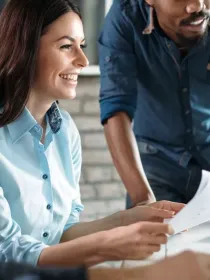 A group of people in a bright office setting, focused on a computer screen. A woman in a light blue shirt is smiling and holding papers, while two men, one in a dark blue shirt and the other in a patterned shirt, look on attentively.