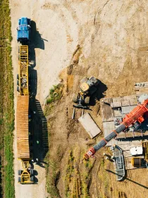 Aerial view of a construction site with a blue truck and yellow trailer on a dirt road next to a green field. A red crane and various construction equipment are on a wooden platform on the right side, with tire tracks visible in the dirt.