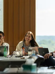 A group of people sit around a table in a meeting room with laptops and water bottles. A man and a woman in the foreground are smiling, while another woman types on a laptop. A man wearing a mask sits nearby. Sunlight filters through the window blinds.
