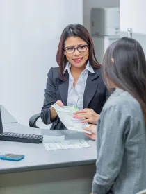 A woman in a business suit and glasses is sitting at a desk, smiling and holding a document. Another person is seated across from her, partially visible, also holding the document. A keyboard and a calculator are on the desk.