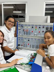 Three young women in white shirts work together at an electrical engineering lab station with equipment and wiring. They are smiling and appear engaged with their project. A notebook and a green folder are on the table.