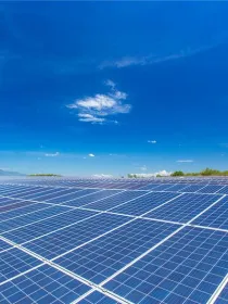 A large field of solar panels under a clear blue sky with a few clouds. In the background, there are distant mountains and green trees.