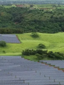 Aerial view of a large solar panel farm on a green hillside, surrounded by lush vegetation and trees. The solar panels are arranged in neat rows, reflecting light. In the background, more green hills and a distant landscape are visible.