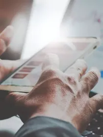 Close-up of a person using a smartphone with financial charts displayed on a laptop screen in the background.