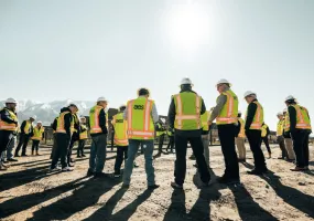 A group of people wearing hard hats and high-visibility vests stand in a circle on a construction site with mountains in the background. The sun shines brightly in the clear sky.