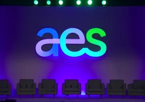 A stage setup with the large letters 'aes' illuminated in blue and green against a purple backdrop. Six empty gray chairs are lined up in front of the backdrop, and a podium with a microphone is on the right side.