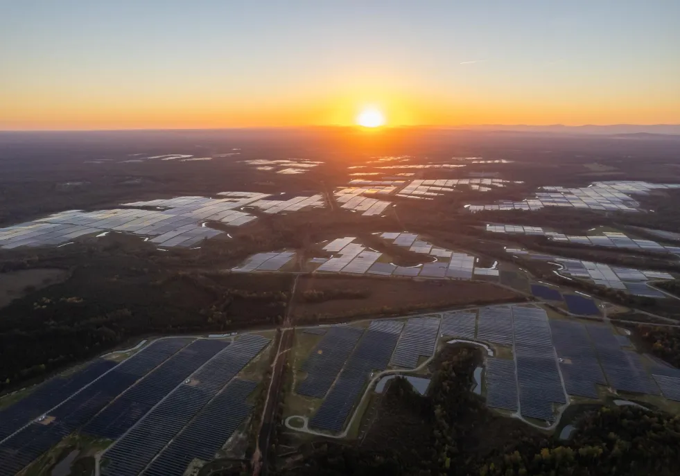 Aerial view of a vast solar farm at sunset, with rows of solar panels reflecting the sun's orange glow. The landscape is surrounded by forests and distant mountains under a clear sky.