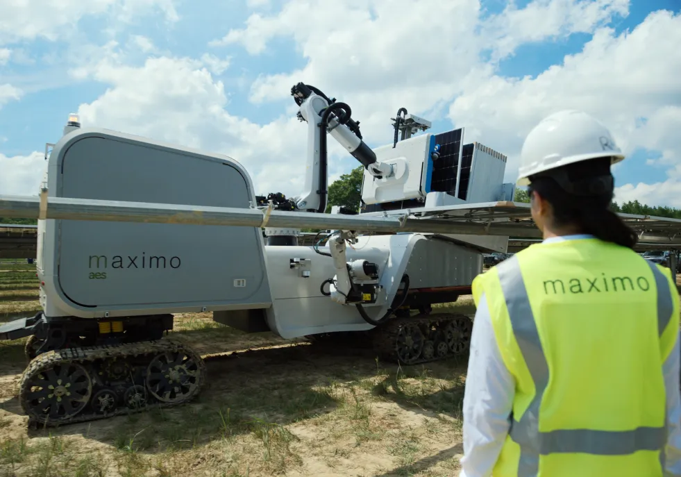 A person wearing a hard hat and a vest labeled 'maximo' stands in front of a large robotic machine on tracks. The machine has a mechanical arm and is situated outdoors under a partly cloudy sky.