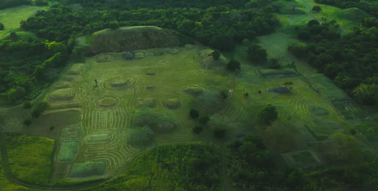Aerial view of an ancient archaeological site with circular and rectangular mounds surrounded by lush green vegetation.