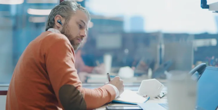 A man with a beard wearing an orange sweater is sitting at a desk, writing in a notebook. He is wearing earbuds and looking intently at his work. A laptop, smartphone, and a face mask are on the desk. The background shows a blurred office setting with large windows.