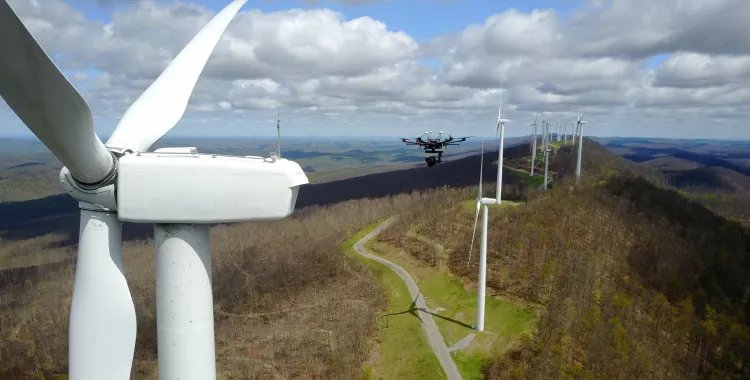 Aerial view of a wind farm with several wind turbines on a hilly landscape. A drone is flying near one of the turbines under a cloudy sky.