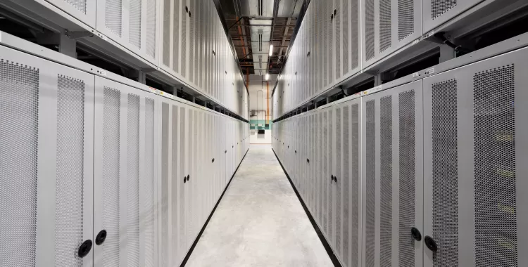 A narrow corridor in a data center with rows of tall, white server cabinets on both sides. The cabinets have perforated doors and black handles. The corridor has a concrete floor and overhead lighting. The perspective leads to a distant wall with visible pipes and panels.