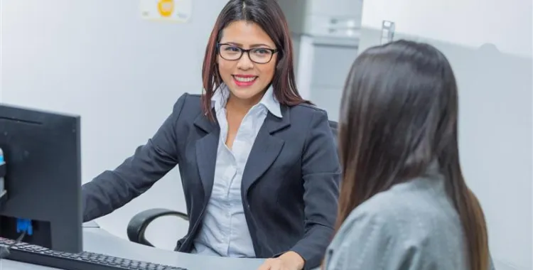 A woman in a business suit and glasses smiles while sitting at a desk with a computer, talking to another person whose back is to the camera.