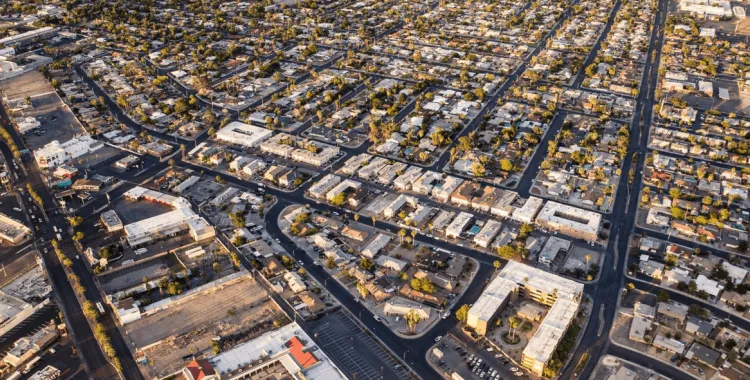 Aerial view of a densely populated urban area with a grid-like street layout. The streets are lined with residential buildings, small commercial properties, and scattered trees. The buildings have flat roofs and are closely packed together, creating a uniform pattern. The image is taken during daylight, casting shadows and highlighting the orderly arrangement of the neighborhood.