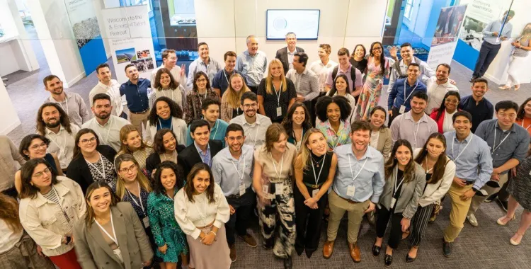 A large group of diverse professionals smiling and posing for a group photo at an indoor corporate event, with banners and screens in the background indicating a retreat or conference setting.