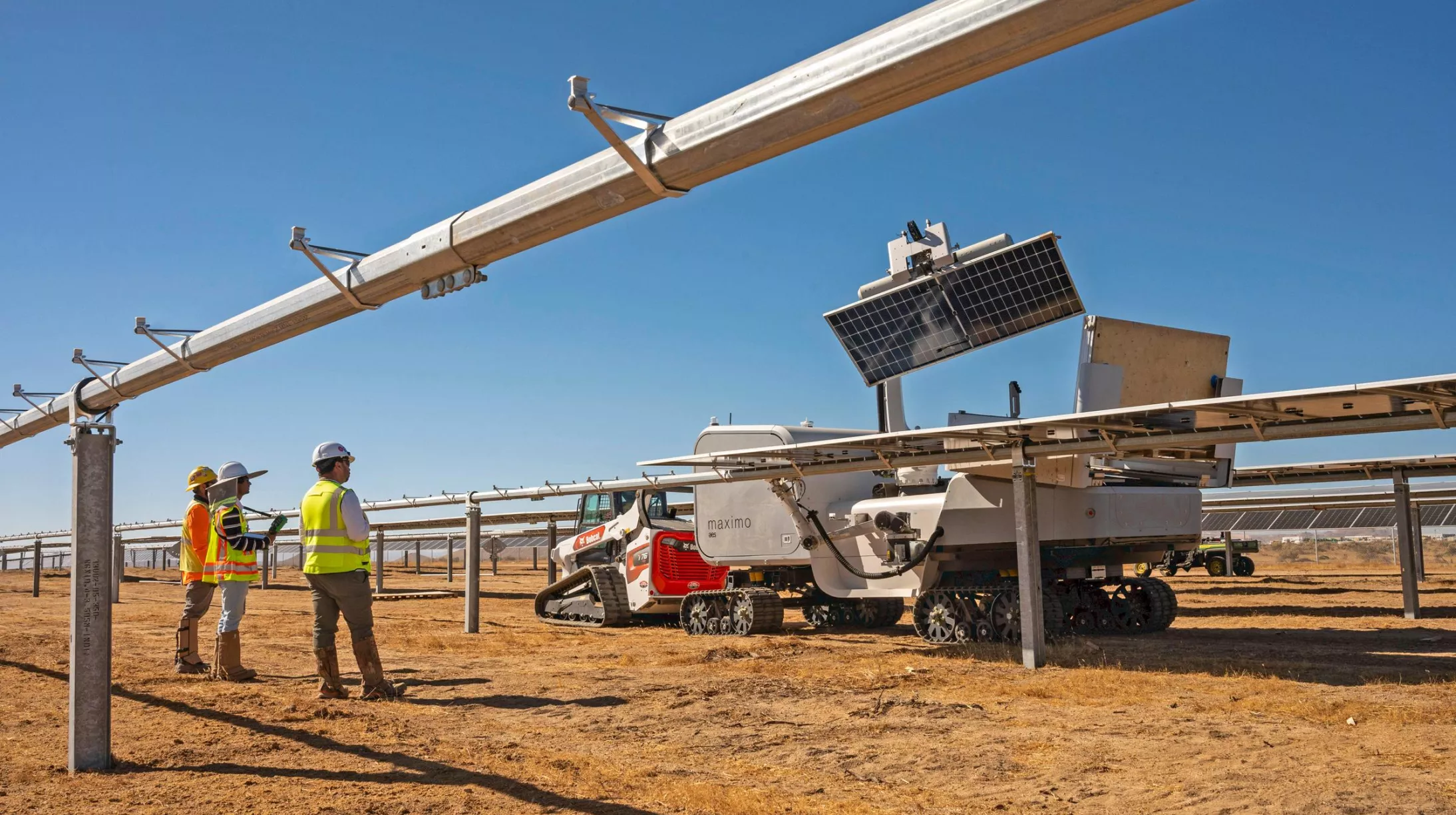 Three workers in safety gear stand beside a solar panel installation in a dry, open field. A robotic machine with tracks is positioned under the panels, likely for maintenance or cleaning. The sky is clear and blue.