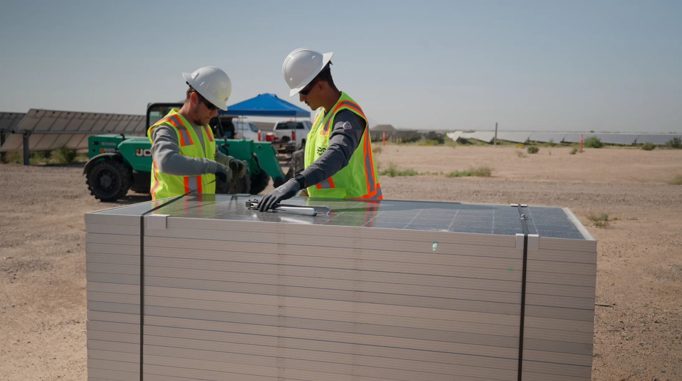 Two workers wearing safety vests and helmets are inspecting a stack of solar panels at a construction site. They are in a desert-like area with solar panels and machinery in the background.