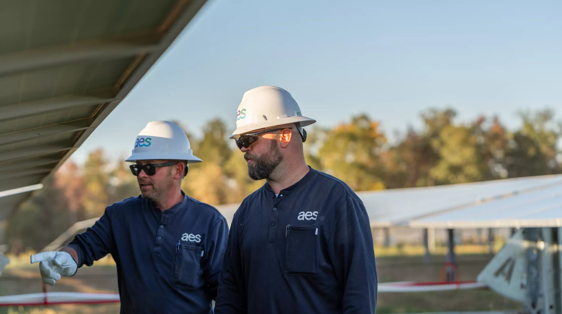 Two workers wearing AES uniforms and hard hats are inspecting solar panels in a field. One worker is pointing at the panels. Trees are visible in the background under a clear sky.