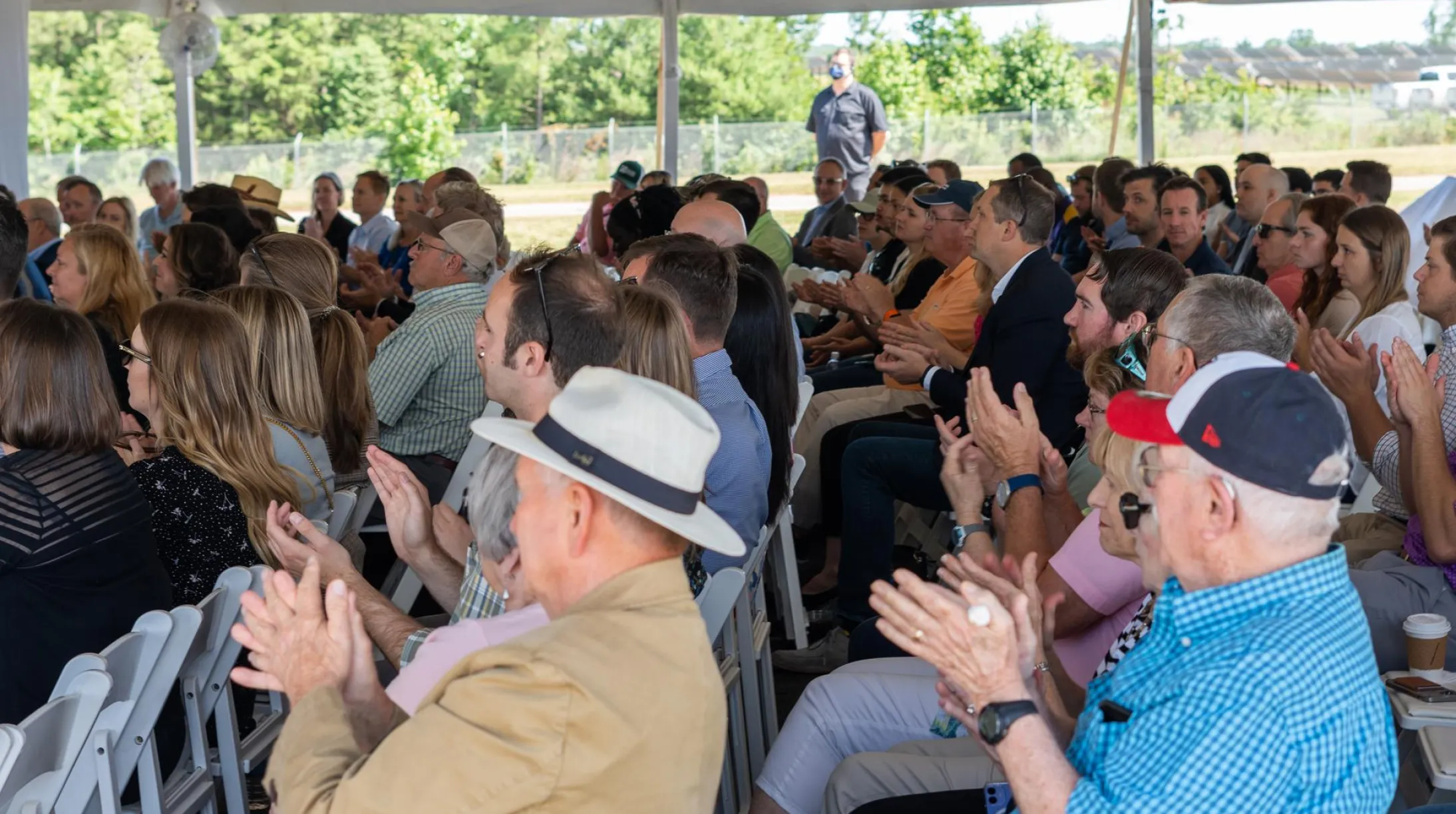 A large group of people sitting under a tent, clapping and facing forward. The audience includes men and women of various ages, some wearing hats. The setting is outdoors with trees visible in the background.