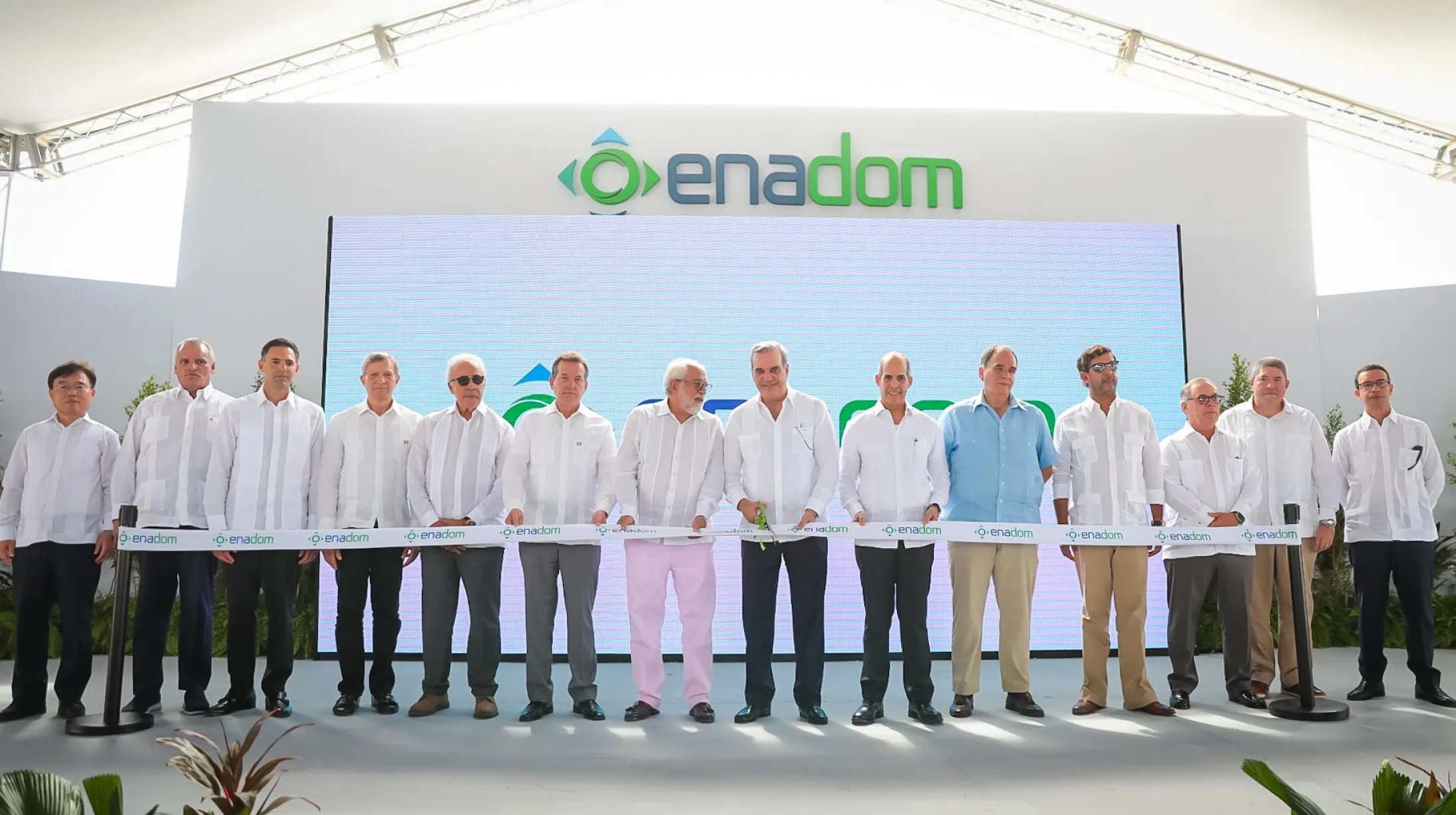 A group of men in white shirts stand in a line holding a ribbon with 'enadom' printed on it, in front of a large sign with the same logo. The setting appears to be a formal event under a tent.