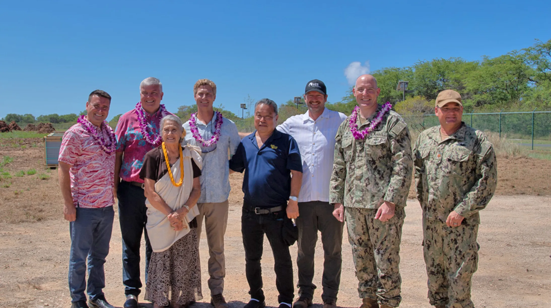 A group of eight people stands outdoors on a sunny day. Some are wearing military uniforms, while others wear casual or Hawaiian-style clothing with leis around their necks. They are standing on a dirt path with trees and a clear blue sky in the background.