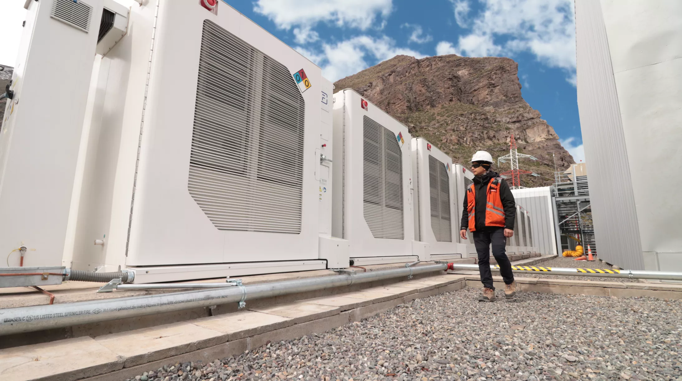 A person wearing a hard hat and orange safety vest walks beside large industrial cooling units in a mountainous area. The sky is clear with a few clouds.