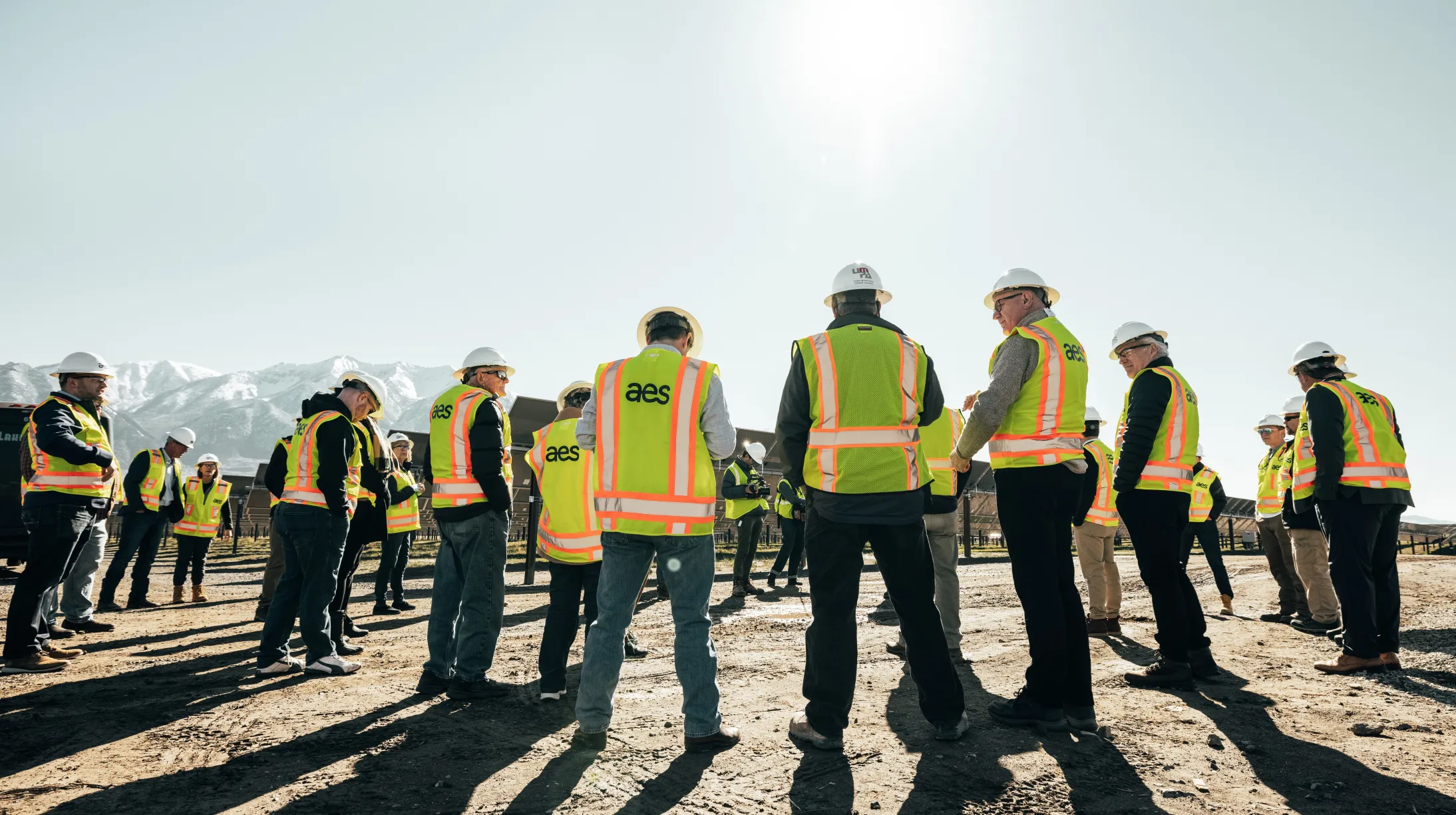 A group of people wearing yellow safety vests and hard hats stand outside on a sunny day. They are gathered on a dirt surface with mountains visible in the background.