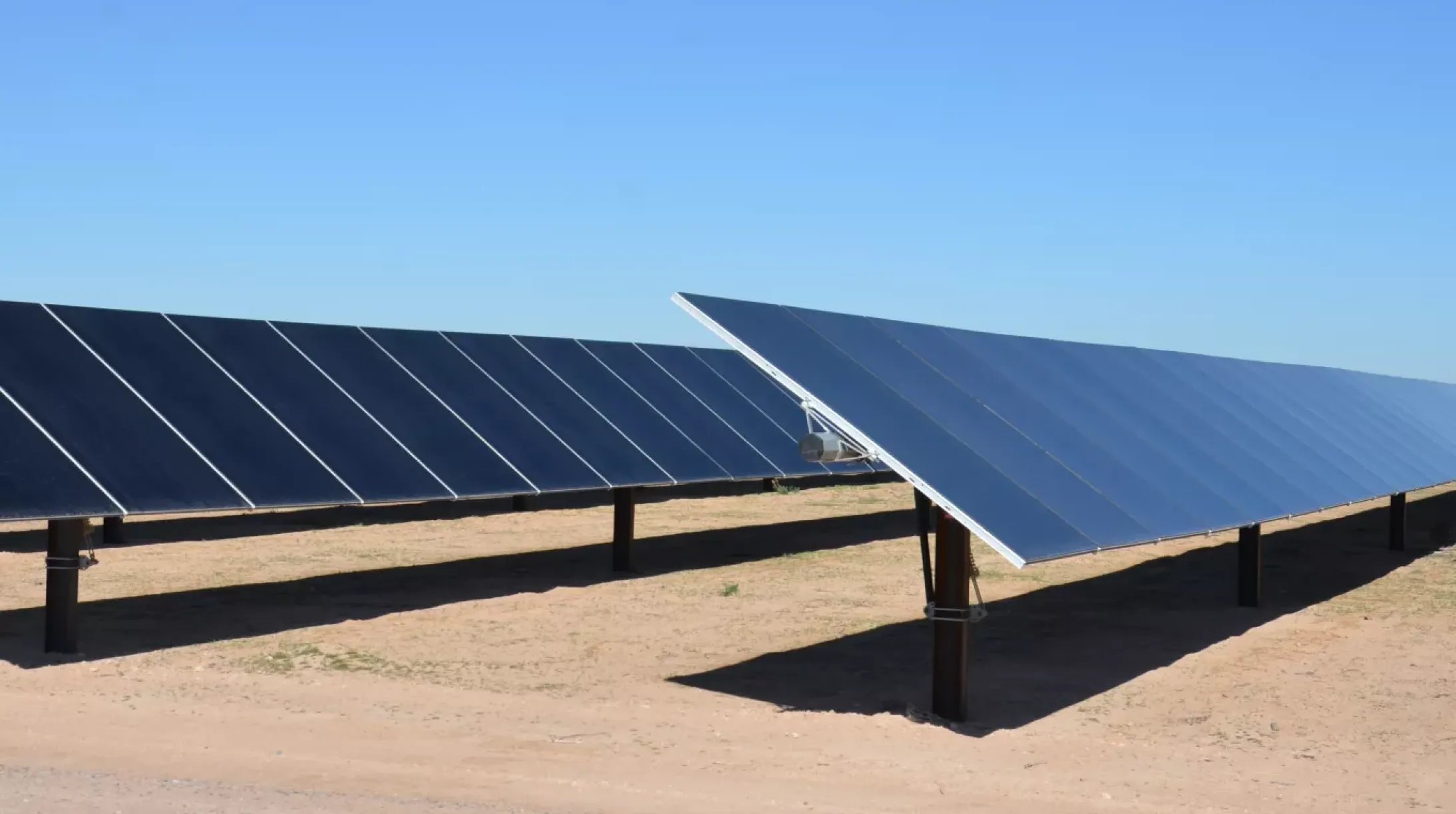 Rows of solar panels on a sandy ground under a clear blue sky.