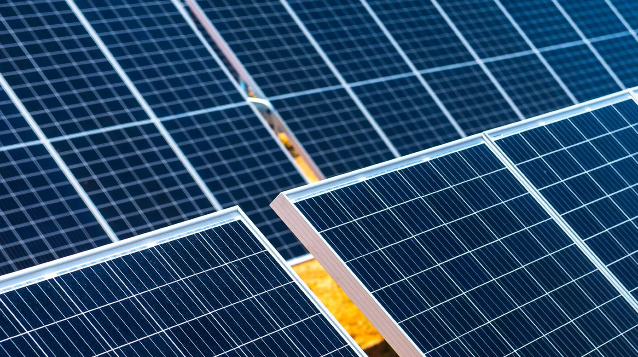 Close-up view of multiple rows of solar panels arranged in a grid pattern, reflecting sunlight. The panels are set on a grassy field, with some green patches visible between the rows.