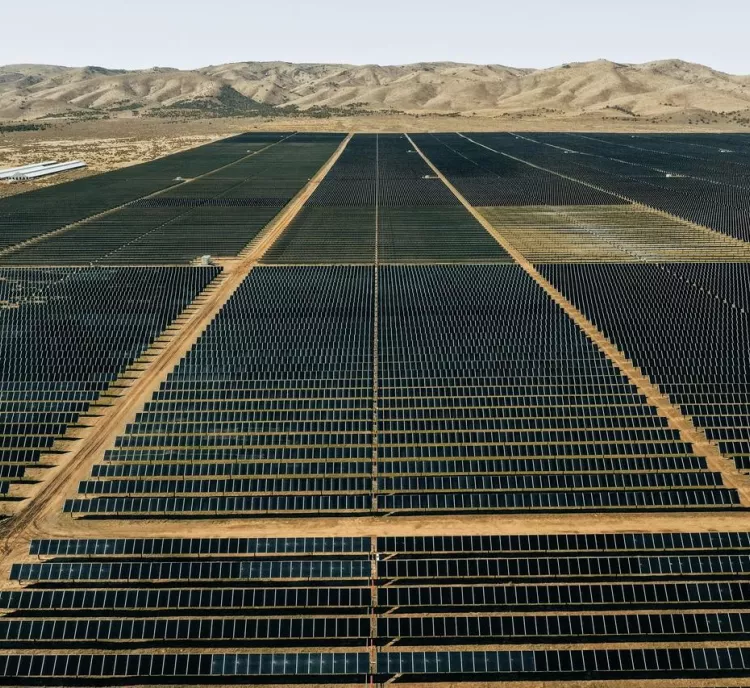 Aerial view of a vast solar farm with rows of solar panels stretching into the distance. The panels are arranged in a grid pattern on a flat, arid landscape. In the background, there are low, barren hills under a clear sky.