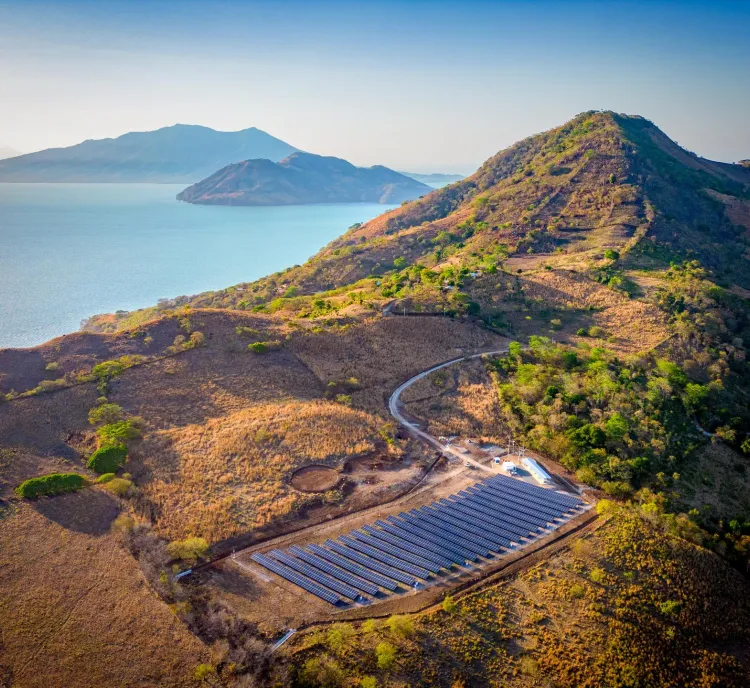 Aerial view of a solar panel farm situated on a hilly landscape near a large body of water. The hills are covered with sparse vegetation and the water reflects a clear blue sky. A winding dirt road leads to the solar panels, surrounded by patches of green trees and brown grass.