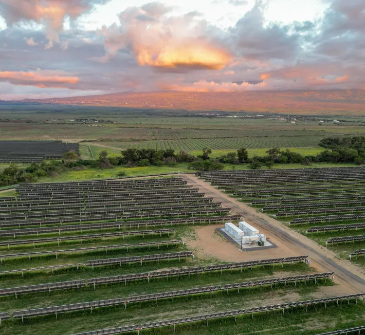 Aerial view of a large solar farm with rows of solar panels stretching across a green landscape. In the background, a mountain range is visible under a sky filled with colorful clouds at sunset.