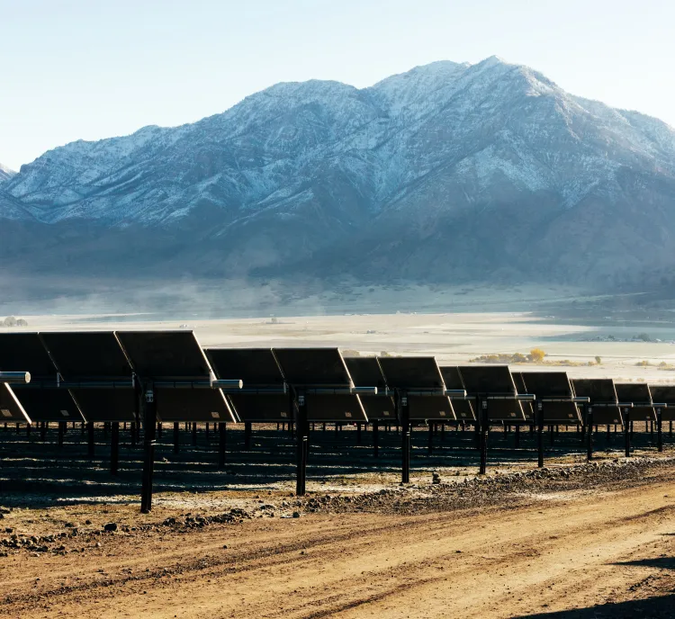 Rows of solar panels are installed on a dirt field, with a backdrop of snow-capped mountains under a clear blue sky.