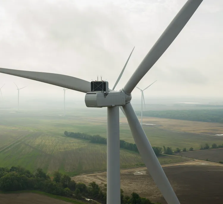 Aerial view of a large wind turbine in the foreground with several other turbines scattered across a vast, flat landscape of fields and trees under a cloudy sky.