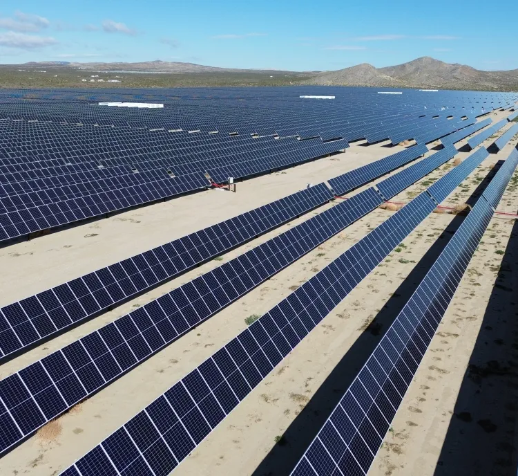 A vast solar farm with numerous rows of blue solar panels stretching into the distance under a clear blue sky. The landscape is arid with mountains visible on the horizon.