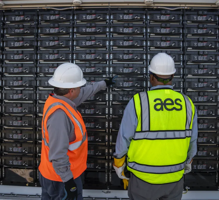 Two workers wearing hard hats and safety vests stand in front of a large battery storage system. One worker is pointing towards the batteries, while the other observes. The vest of the worker on the right has 'aes' written on it.