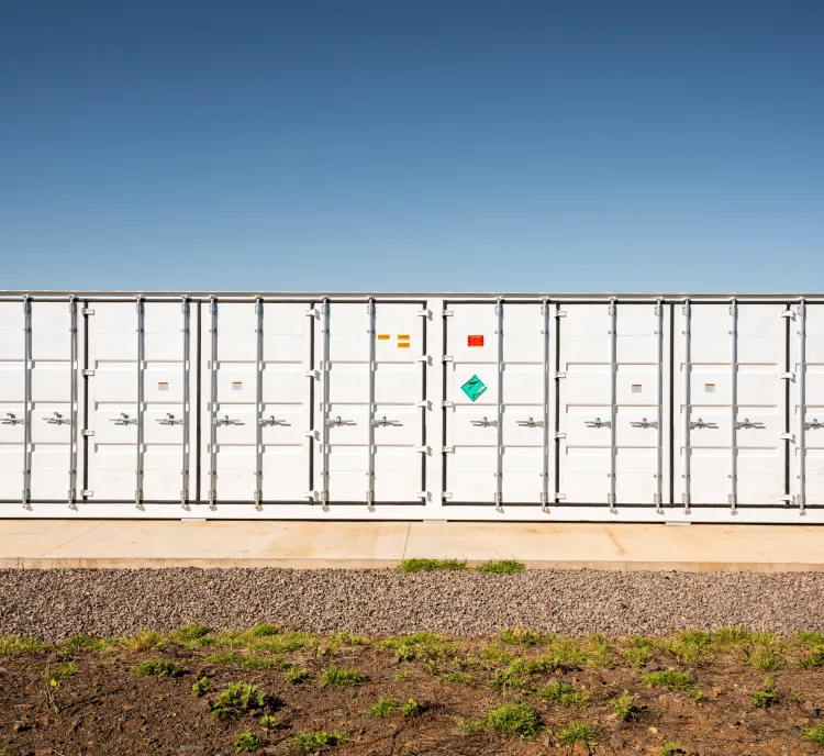 A large white industrial container with multiple doors and hazard labels is situated on a concrete platform. The background shows a clear blue sky and part of a solar panel array on the left. The foreground has gravel and patches of grass.