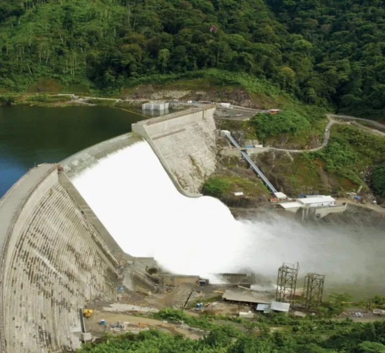 Aerial view of a large dam with water flowing over its spillway, surrounded by lush green forest. The dam is curved and made of concrete. A mist forms at the base where the water impacts. Roads and small buildings are visible near the dam.