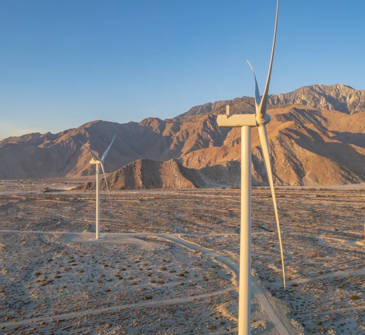 Aerial view of wind turbines in a desert landscape with mountains in the background. The turbines are surrounded by sparse vegetation under a clear blue sky during sunset, casting long shadows on the ground.