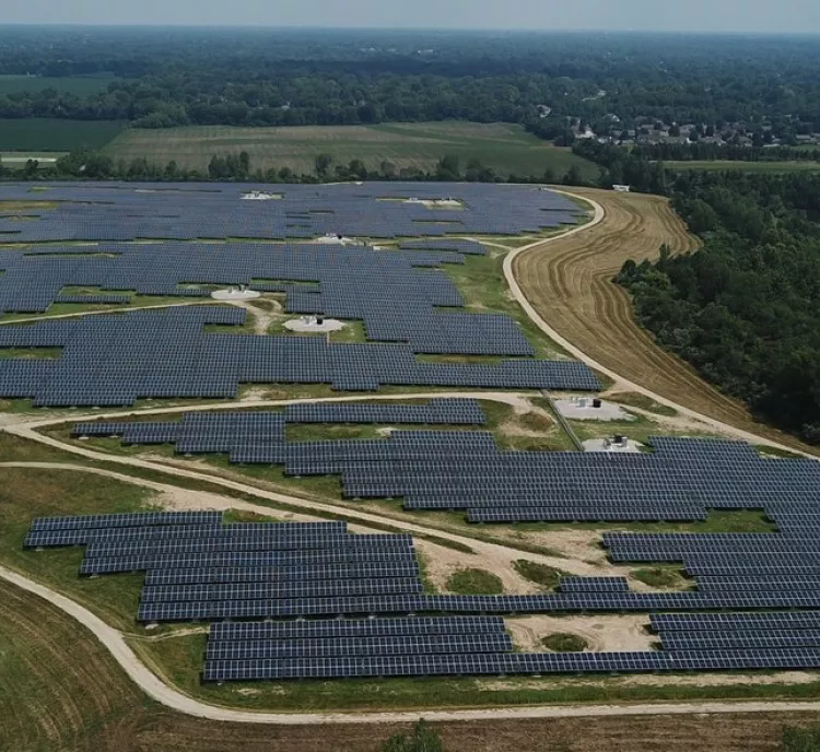 Aerial view of a large solar panel farm with rows of solar panels covering open fields. A small building and several parked cars are visible in the foreground. The landscape includes green fields, a river, and distant trees under a clear sky.