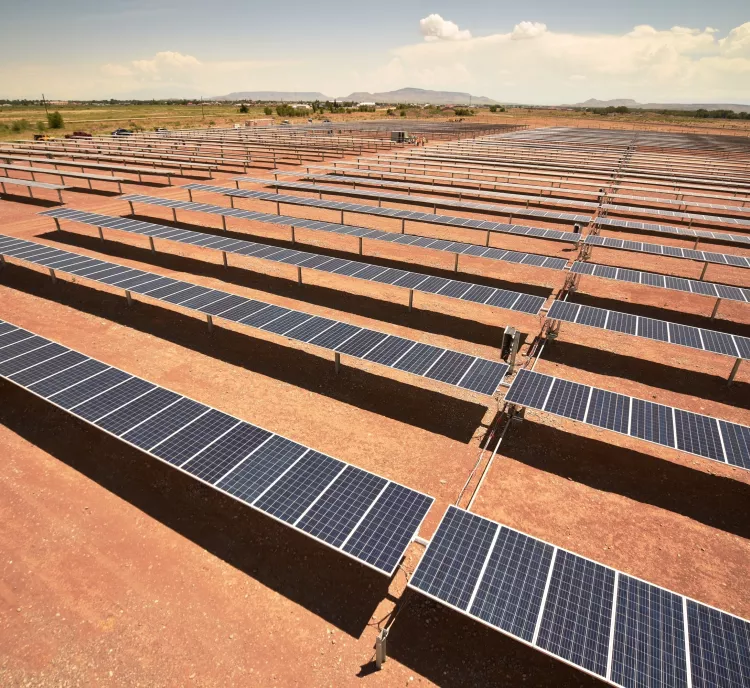 A large solar farm with rows of solar panels installed on a barren landscape. The panels are aligned in parallel rows under a clear blue sky. In the background, there is a view of distant mountains and some greenery.