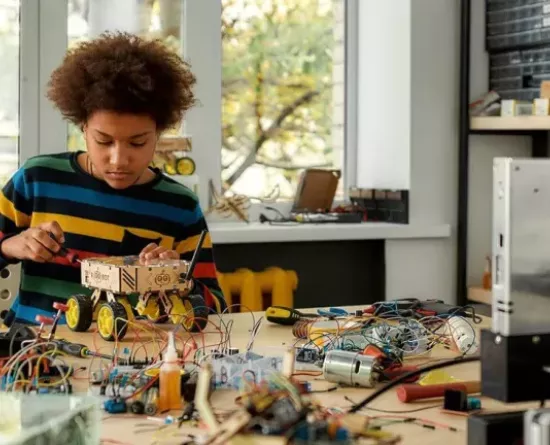 A young person wearing a striped sweater works on assembling a small robotic vehicle at a cluttered desk filled with electronic components and wires. The room has large windows with a view of trees outside. Shelves filled with tools and parts are visible in the background.