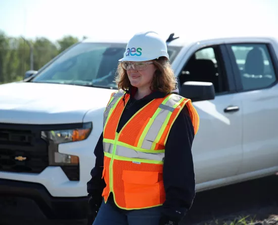 A person wearing a white hard hat with the AES logo, safety goggles, and an orange high-visibility vest stands in front of a white pickup truck. The background shows a blurred outdoor setting with trees.