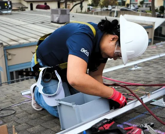 A worker wearing a white hard hat, safety glasses, and a blue shirt with a logo is installing equipment on a rooftop. They are kneeling next to a gray box and using tools. Safety harnesses and ropes are visible, ensuring secure positioning on the roof. Buildings and trees are in the background.