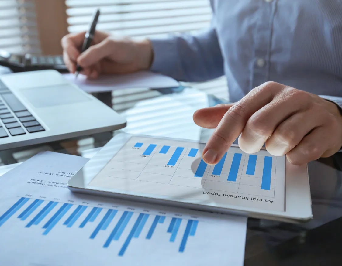 A person in a blue shirt is analyzing data on a tablet showing a bar chart. They are pointing at the screen with one hand while writing on paper with the other. A laptop and additional paper with charts are on the glass table.