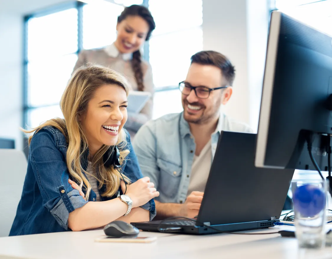 Three people in an office setting are smiling and looking at a computer screen. A woman in the foreground is laughing, with headphones around her neck. A man next to her is also smiling, and another woman stands behind them, holding a tablet. The environment is bright with natural light coming through large windows.