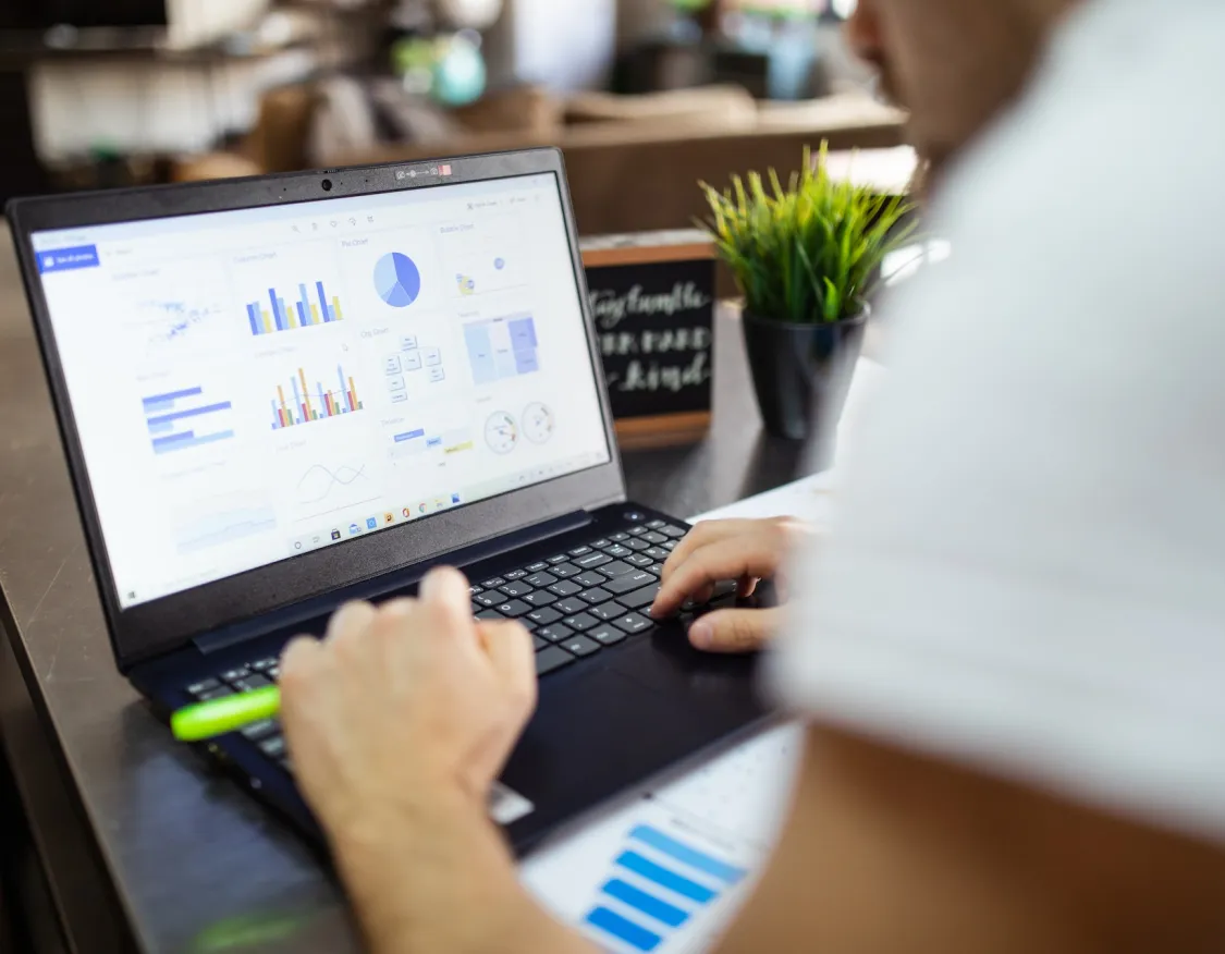 A person using a laptop displaying various charts and graphs, including bar, pie, and line charts. The individual is holding a green pen and working at a desk with a small potted plant nearby.