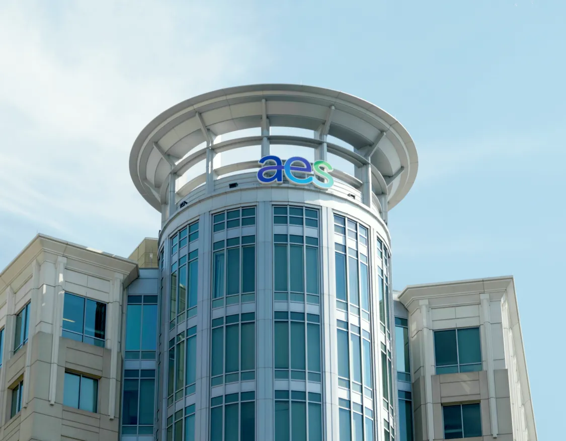 Modern building with a cylindrical glass tower and the letters 'aes' on top against a clear blue sky.